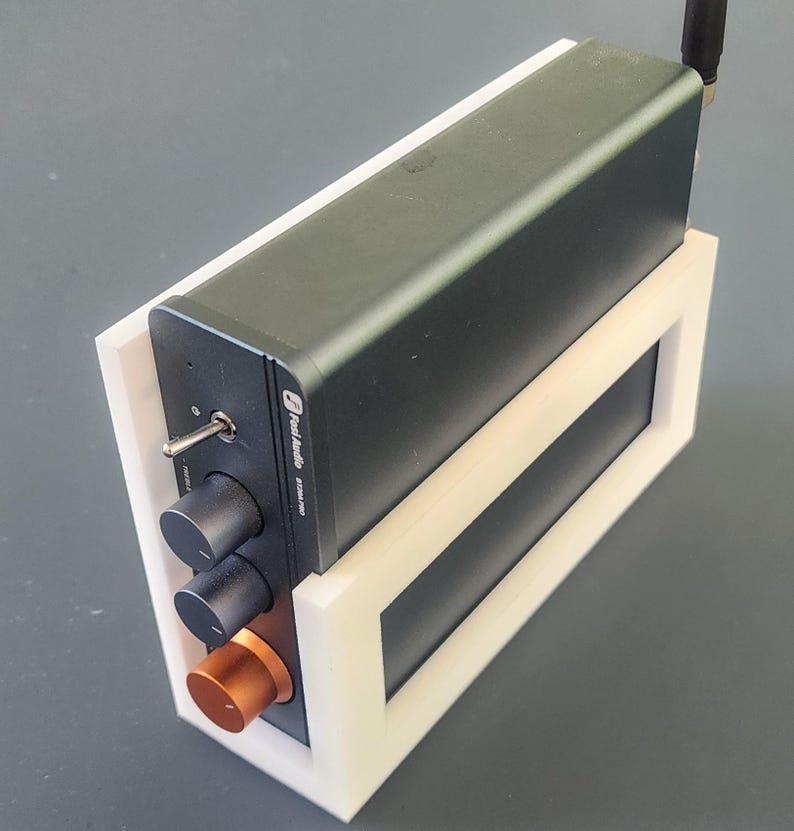 May include: A black audio amplifier with a silver toggle switch, two black knobs, and a copper-colored knob. The amplifier is mounted in a white frame and has a small antenna. The text "Fosi Audio BT30A PRO" is visible on the device.