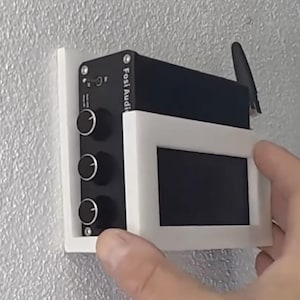 May include: A black electronic device with three knobs and a small screen, mounted on a white bracket against a textured wall. The device has the text "Fosi Audio" printed on it. An antenna is attached to the top.