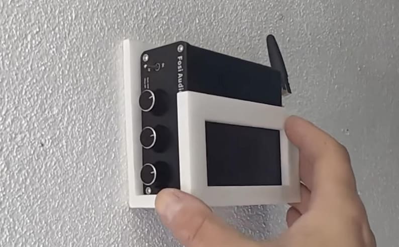 May include: A black electronic device with three knobs and a small screen, mounted on a white bracket against a textured wall. The device has the text "Fosi Audio" printed on it. An antenna is attached to the top.