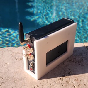 May include: A white and black electronic device with a black antenna and various connection ports. The device has a rectangular black screen and is set against a backdrop of a pool.