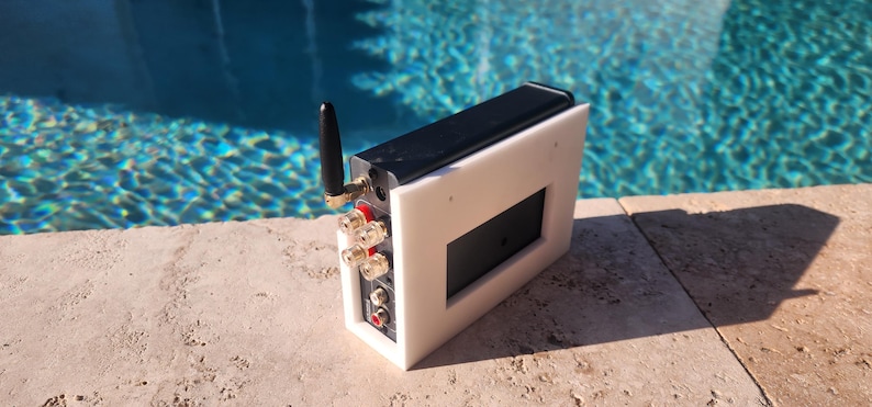 May include: A white and black electronic device with a black antenna and various connection ports. The device has a rectangular black screen and is set against a backdrop of a pool.
