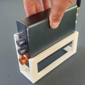 May include: A dark gray electronic device with knobs and a copper-colored accent, held in a white 3D-printed stand. The device has a power switch and multiple ports. The stand is rectangular with an open design.
