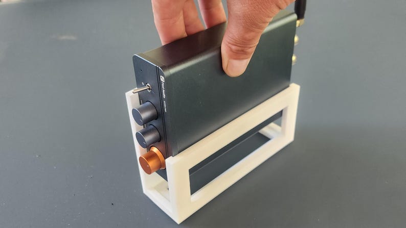 May include: A dark gray electronic device with knobs and a copper-colored accent, held in a white 3D-printed stand. The device has a power switch and multiple ports. The stand is rectangular with an open design.