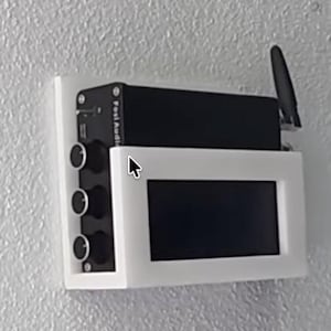 May include: A black electronic device with knobs and a screen, mounted on a white frame. The device has an antenna and the text "Fosi Audio" is visible. The setup is mounted on a textured white wall.
