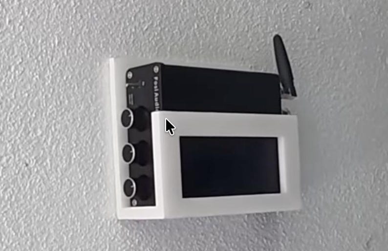 May include: A black electronic device with knobs and a screen, mounted on a white frame. The device has an antenna and the text "Fosi Audio" is visible. The setup is mounted on a textured white wall.