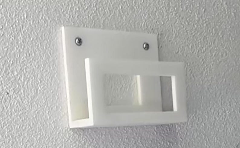 May include: A white, wall-mounted shelf with a rectangular backplate and a projecting, open-fronted shelf. Two silver screws secure the shelf to the wall. The shelf is made of a solid, opaque material.