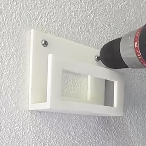 May include: A white, rectangular wall-mounted shelf is being installed with a black and red Craftsman drill. The shelf has a rectangular cutout and is attached to a textured, light gray wall with screws.