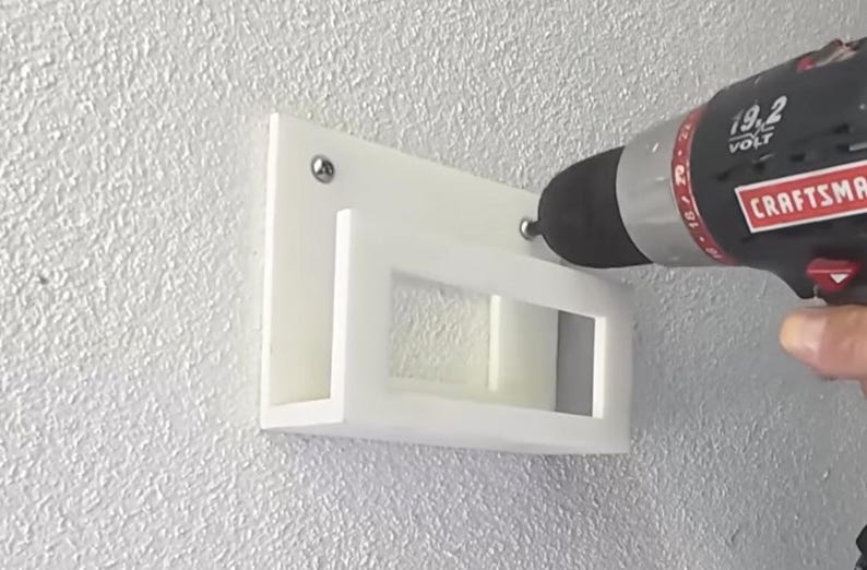 May include: A white, rectangular wall-mounted shelf is being installed with a black and red Craftsman drill. The shelf has a rectangular cutout and is attached to a textured, light gray wall with screws.