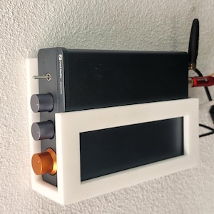 May include: A black audio amplifier with silver knobs and an orange knob, mounted in a white frame. The amplifier has a small antenna and is labeled "Post Audio". The frame is mounted on a textured wall.