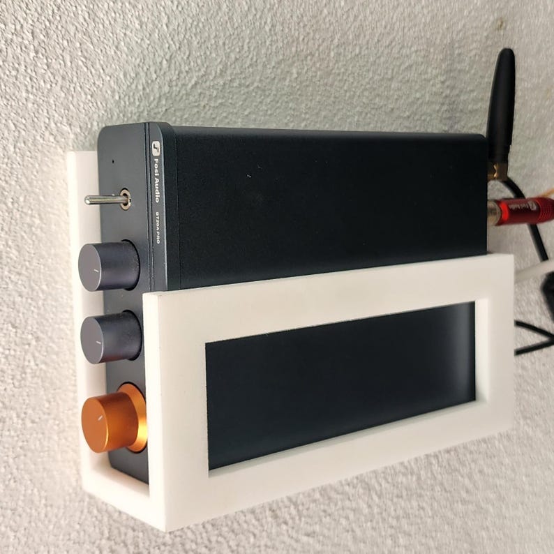May include: A black audio amplifier with silver knobs and an orange knob, mounted in a white frame. The amplifier has a small antenna and is labeled "Post Audio". The frame is mounted on a textured wall.