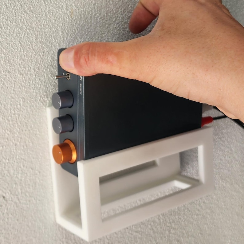 May include: A black audio device with silver knobs and an orange knob is mounted on a white rectangular shelf. The device is being held by a hand. The shelf is mounted on a textured white wall.