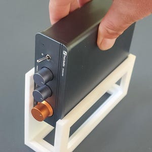 May include: A black audio amplifier with silver knobs and an orange volume control, held in a white stand. The amplifier has the text "Fosi Audio BT20A Pro".