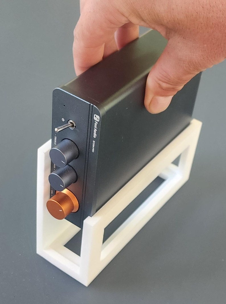 May include: A black audio amplifier with silver knobs and an orange volume control, held in a white stand. The amplifier has the text "Fosi Audio BT20A Pro".