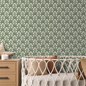 Pode incluir: Papel de parede verde sálvia com um padrão floral branco repetido. Uma cômoda de madeira castanho claro com puxadores pretos está em primeiro plano, junto com um berço branco. Os itens decorativos incluem um elefante de pelúcia e uma almofada em forma de estrela.