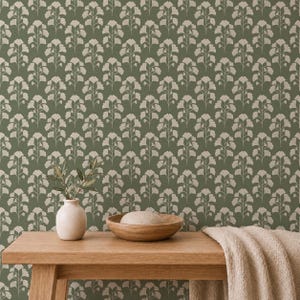 May include: A green and beige floral wallpaper pattern. A wooden table holds a small white vase with greenery, a wooden bowl with a stone, and a beige folded blanket. The wallpaper has a repeating floral design.
