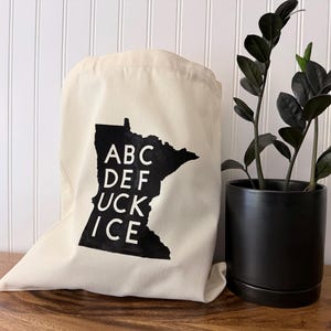 May include: A cream-colored tote bag with a black graphic of the state of Minnesota and the words "ABC DEF UCK ICE" in white. A black potted plant is to the right of the bag, set against a white paneled wall.