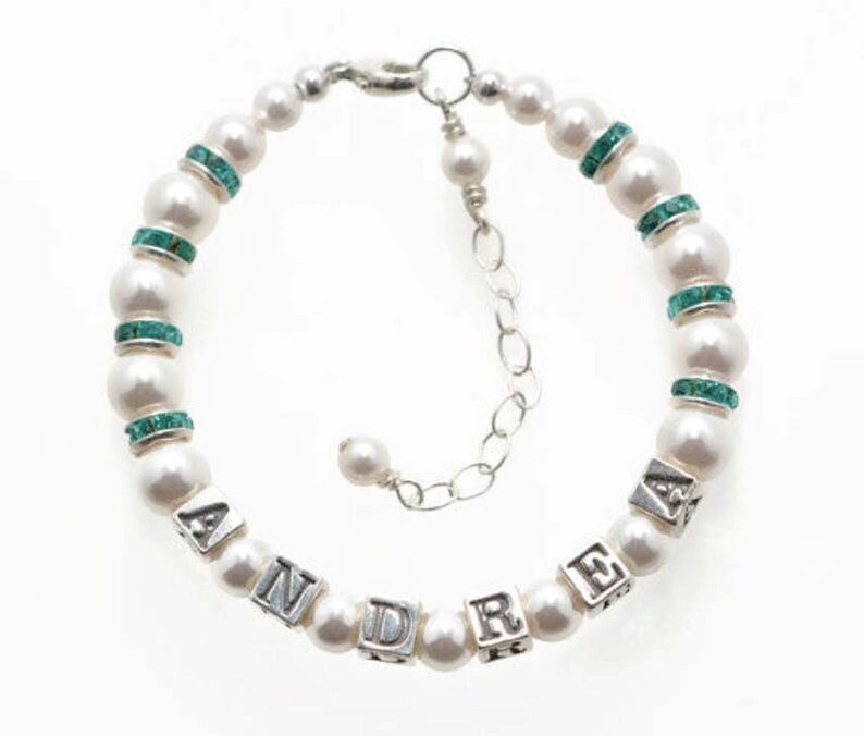 May include: A pearl bracelet with the name "ANDRE" spelled out in silver letter beads. The bracelet features white pearls, interspersed with turquoise-colored rhinestone spacers, and a silver chain clasp.