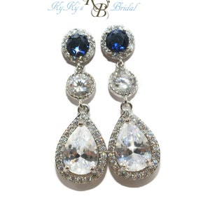 May include: A pair of elegant drop earrings with a three-tiered design. The top features a dark blue, round stone surrounded by smaller clear stones. The middle tier has a clear, round stone, and the bottom is a clear, pear-shaped stone, also surrounded by clear stones. The earrings are silver-toned.