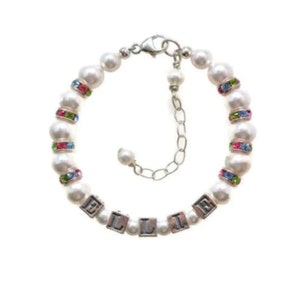 May include: A white pearl bracelet with the name "ELLIE" spelled out in silver letter beads. The bracelet features colorful rhinestone accents and a silver clasp. The bracelet is designed for a child.