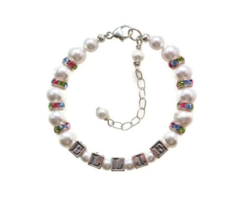 May include: A white pearl bracelet with the name "ELLIE" spelled out in silver letter beads. The bracelet features colorful rhinestone accents and a silver clasp. The bracelet is designed for a child.