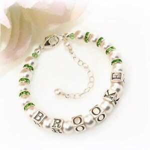 May include: A pearl bracelet with silver accents. The bracelet features white pearls, green rhinestone spacers, and silver lettered cubes spelling out a name. The clasp and extender chain are also silver.