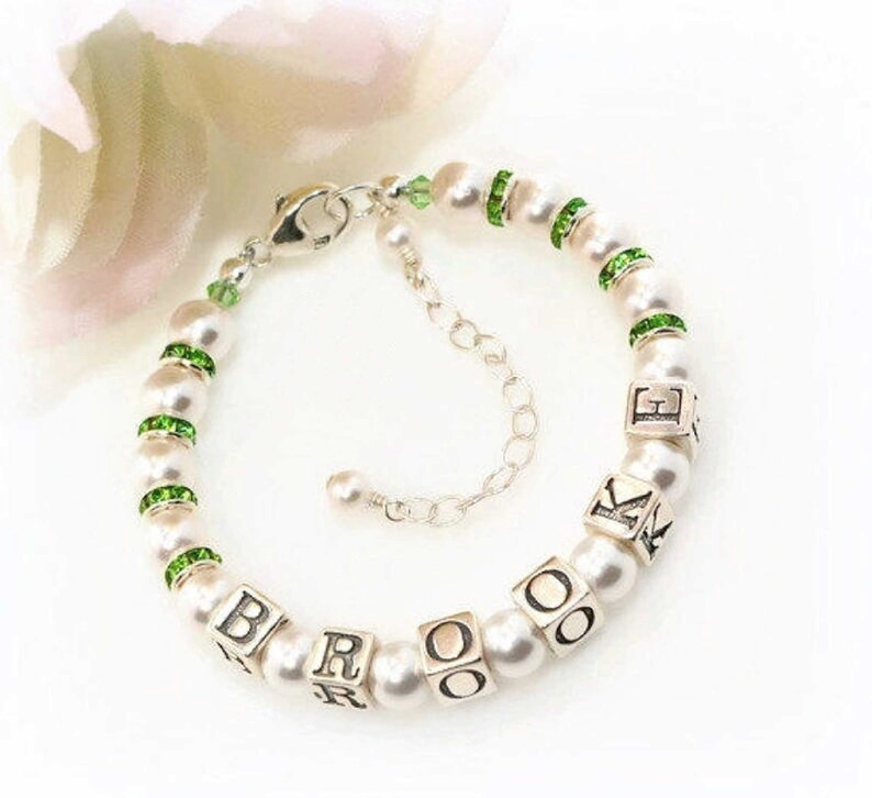 May include: A pearl bracelet with silver accents. The bracelet features white pearls, green rhinestone spacers, and silver lettered cubes spelling out a name. The clasp and extender chain are also silver.