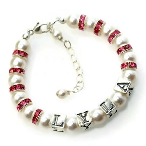 May include: A pearl bracelet with pink rhinestone accents and silver letter beads spelling "LYLA". The bracelet features a silver clasp and an adjustable chain. The pearls are white and the rhinestones are a vibrant pink.