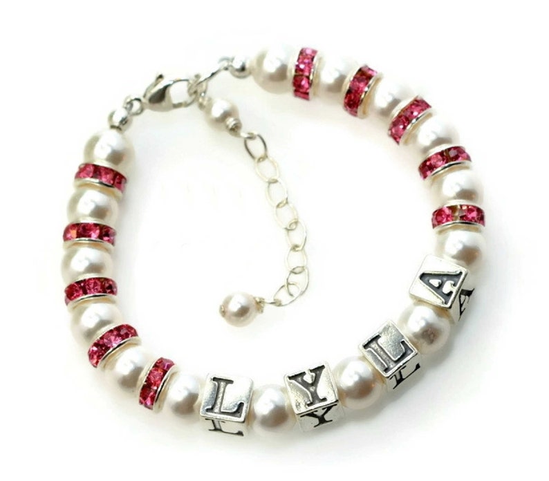 May include: A pearl bracelet with pink rhinestone accents and silver letter beads spelling "LYLA". The bracelet features a silver clasp and an adjustable chain. The pearls are white and the rhinestones are a vibrant pink.
