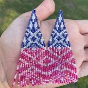 Puede incluir: Un par de pendientes de cuentas con un diseño patriótico. La parte superior presenta un patrón geométrico azul y blanco, que se transforma en una franja rosa y blanca. Los pendientes son triangulares y se sostienen en una mano.