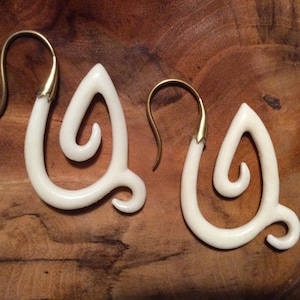 May include: A pair of cream-colored, teardrop-shaped earrings with brass-colored hooks. The earrings have a unique, swirling design. The earrings are displayed on a wooden surface.