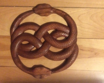 May include: A wooden sculpture of two snakes intertwined in a circle, creating a knot-like design. The snakes are carved with scales and have a dark brown colour.