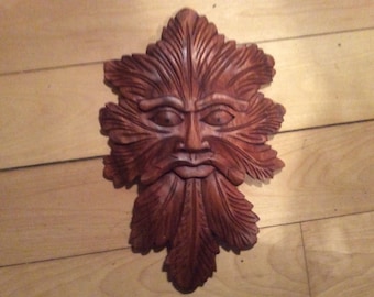 May include: A wooden carving of a Green Man's face, with a leafy beard and a serious expression. The carving is painted in shades of brown and has a rustic, natural look.