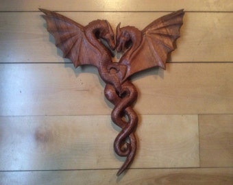 May include: A wooden carving of two dragons intertwined in a heart shape. The dragons are brown and have wings. The carving is on a light wood background.