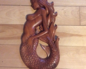 May include: A wooden sculpture of a mermaid with a detailed tail and flowing hair. The sculpture is carved from a light brown wood and has a natural finish.