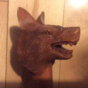 May include: A wooden carving of a wolf's head with an open mouth and sharp teeth. The carving is detailed and realistic, with visible wood grain.