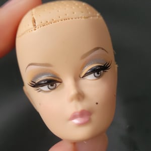 以下が含まれることがあります： 明るい肌色のハゲた人形の頭部。ペイントされたグレーの目、黒いまつげ、アーチ型の茶色の眉毛、ピンクの唇が特徴。頭頂部は髪を取り付けるための質感のある穴あき加工が施されており、頬には小さなほくろがある。