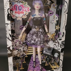 Puede incluir: Una muñeca Barbie Collector Platinum Label con cabello morado y tatuajes visibles en brazos y pecho. La muñeca viste un top negro sin tirantes y una falda estampada, presentada en una caja con la marca 10th Anniversary Tokidoki.