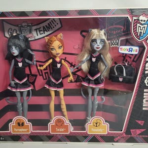 Puede incluir: Un set de Monster High en caja con tres muñecas felinas llamadas Purrsephone, Toralei y Meowlody. Cada muñeca viste un uniforme de animadora negro y rosa. El empaque incluye una bolsa de deporte negra, un gráfico de canasta de baloncesto y el logo de Toys R Us.