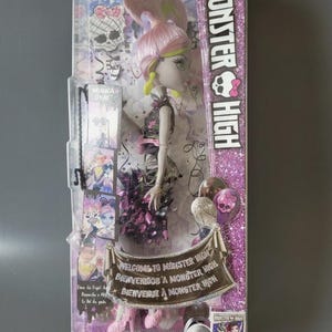 Puede incluir: Una muñeca Monster High en su empaque original con cabello rosa, piel gris y un vestido negro con detalles en rosa. La caja incluye un estandarte que dice Welcome to Monster High, un DVD de 2016 y varias tarjetas de personajes en el lateral.