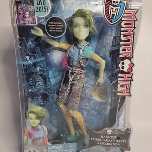 Puede incluir: Una muñeca Monster High Porter Geiss en su empaque original. La muñeca tiene piel verde claro, cabello corto verde y viste una camisa azul con pantalones grises que incluyen un accesorio de cadena. La caja muestra texto como DVD 2015, Haunted y Monster High.