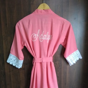 May include: A coral pink robe with white lace trim around the sleeves and hem. The robe has a belt and the name "Adalyn" is embroidered on the back.