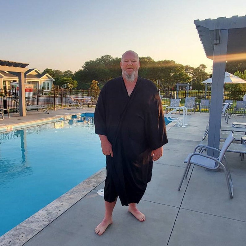 May include: A man wearing a black robe stands by a swimming pool. The pool is surrounded by a gray concrete deck and there are lounge chairs around the pool.