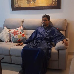May include: A man wearing a dark blue robe sits on a light grey sofa. The sofa has two cushions with floral patterns. The man is looking to the right of the image.