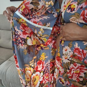 May include: A floral-patterned robe in shades of blue, red, yellow, and pink. The robe features a white trim along the edges and pockets. The fabric is soft and flowing, with a vibrant floral design. The robe is open, revealing the interior lining.