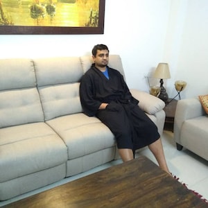 May include: A man wearing a black robe sits on a light gray couch in a living room. The couch is in front of a wall with a framed painting of a seascape. A wooden coffee table sits in front of the couch.