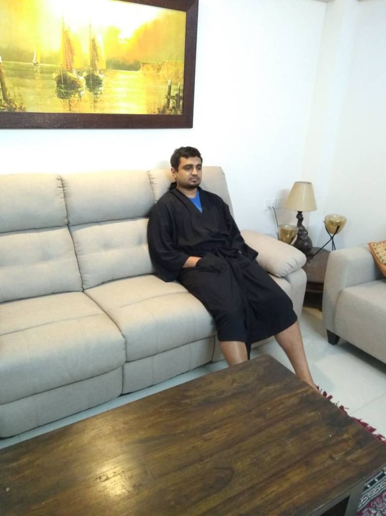 May include: A man wearing a black robe sits on a light gray couch in a living room. The couch is in front of a wall with a framed painting of a seascape. A wooden coffee table sits in front of the couch.