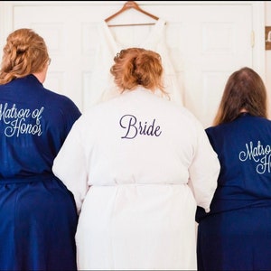 Könnte beinhalten: Drei Personen tragen Roben mit besticktem Text auf der Rückseite. Die Person in der Mitte trägt eine weiße Robe mit dem Wort "Bride" auf der Rückseite. Die Personen an beiden Seiten tragen marineblaue Roben mit dem Text "Matron of Honor" auf der Rückseite.