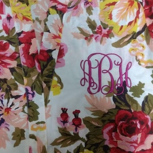 Pode incluir: Um roupão branco com um padrão floral em tons de rosa, vermelho, amarelo e verde. O roupão tem um monograma rosa "ABM" bordado na frente.