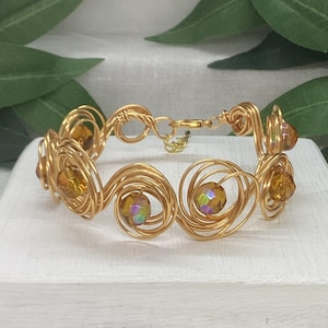 May include: Gold-coloured wire bracelet with swirling patterns and amber beads with iridescent highlights. The bracelet has a delicate clasp and is displayed on a white surface, highlighting its intricate craftsmanship.
