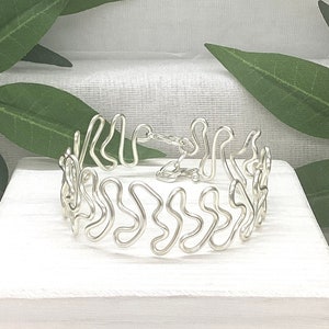 May include: A silver wire bracelet with a distinctive, undulating pattern. The bracelet is composed of interconnected loops and curves, forming an abstract design. The clasp is also silver, securing the bracelet. The jewellery is displayed on a white surface.